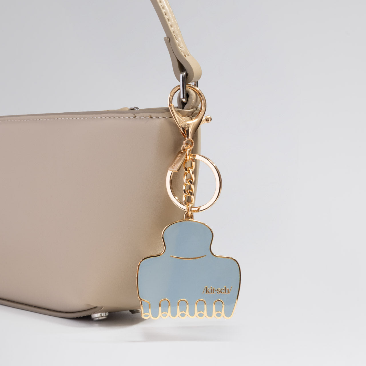 Metal enamel keychain of the kitsch Haze Blue Cloud Clip attached to a beige handbag on a light gray background