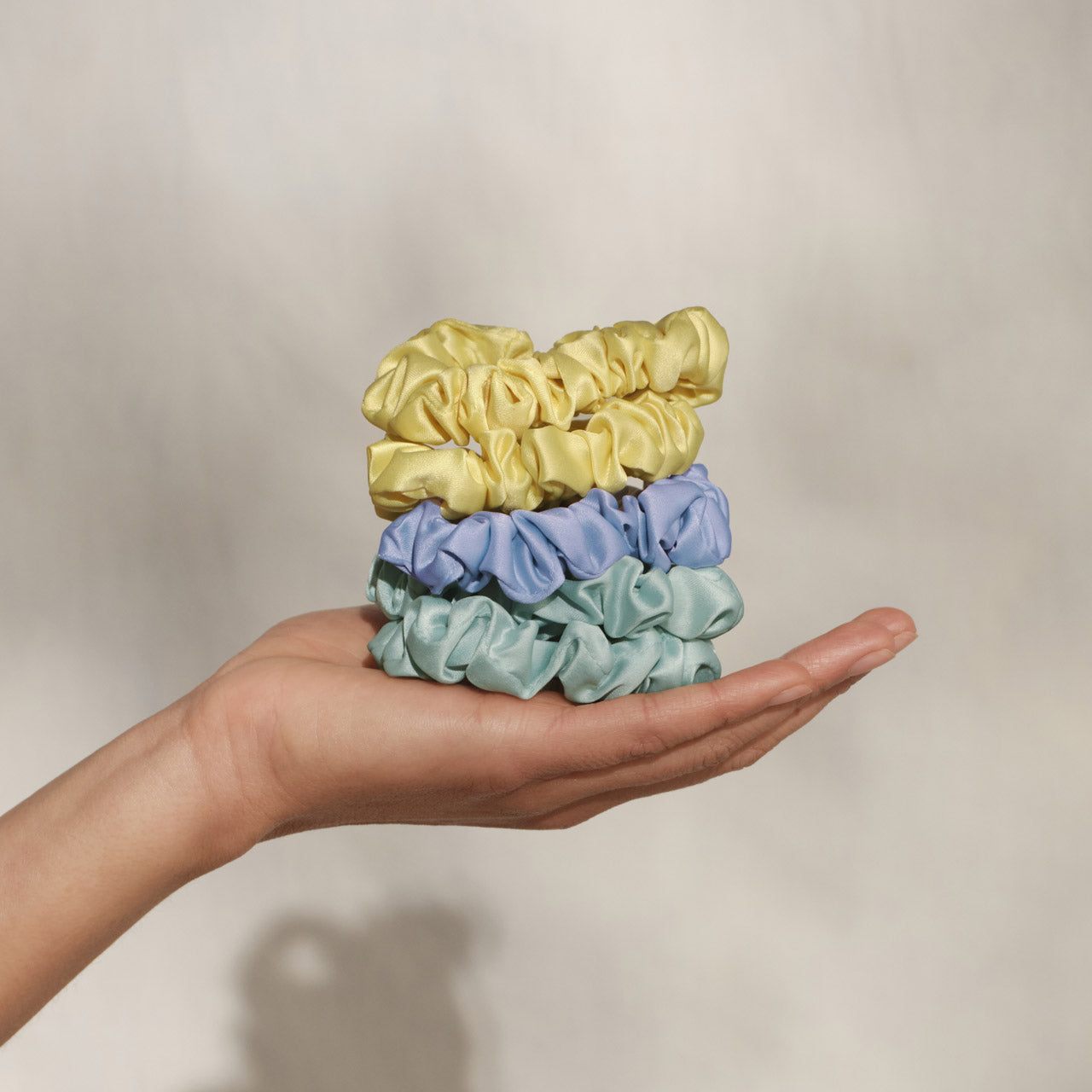Hand holding a stacked set of KITSCH Coastal Cottage Ruched Satin Scrunchies — butter yellow, lavender blue & sage green stacked on palm — product showcase shot — Extra Gentle | 5pcs, silky material, gentle snag-free hold for all hair types
