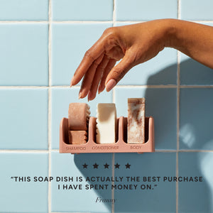 Hand reaching for kitsch shampoo, conditioner, and soap bars in a terracotta soap dish attached to a tiled wall. There is a 5 star customer review on the bottom that reads: This soap dish is actually the best purchase I have spent money on. — Franny